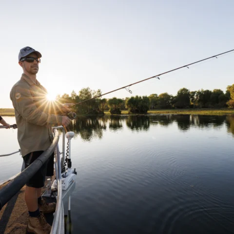 A man in sunglasses and a cap fishes from a boat at dawn, with the sun rising over a calm lake and trees in the background, embracing the beauty of nature.