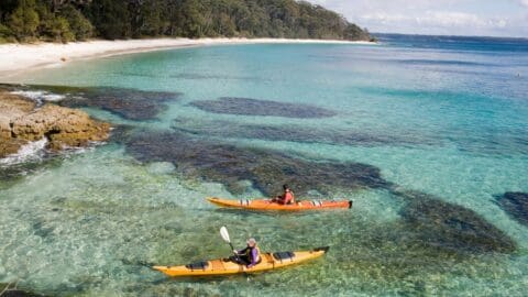 Two people in kayaks paddle on clear turquoise water near a rocky shoreline with a sandy beach and forested area in the background, showcasing the beauty of nature.