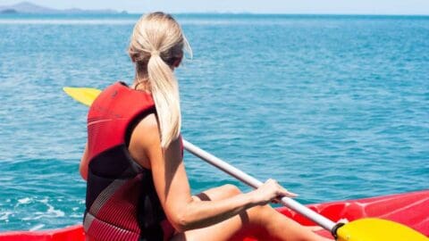 A person with long blonde hair, wearing a red life vest, is kayaking on a large body of water surrounded by nature under a clear blue sky.