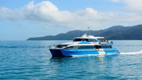 A blue and white boat sails on a calm ocean with a mountainous shoreline and cloudy sky in the background, offering serene views that highlight nature's beauty.