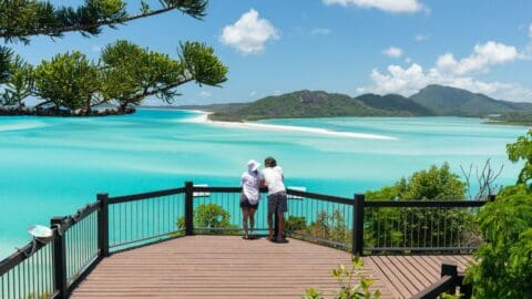 Two people stand on a wooden viewing deck overlooking a turquoise bay surrounded by lush hills under a clear blue sky, embodying the spirit of sustainable ecotourism.