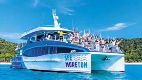A large white and blue boat with "See Moreton" on the side carries a collective of cheering people, set against a backdrop of clear blue water, green trees, and a clear sky.
