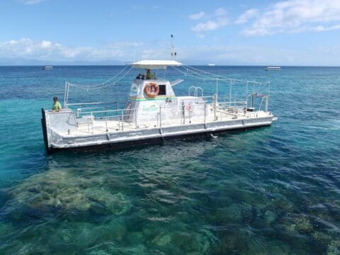 A white boat with a canopy floats on clear, blue-green water near a visible coral reef. A person is standing on the boat. Two other boats are in the background under a partly cloudy sky, offering an eco-friendly tour experience.