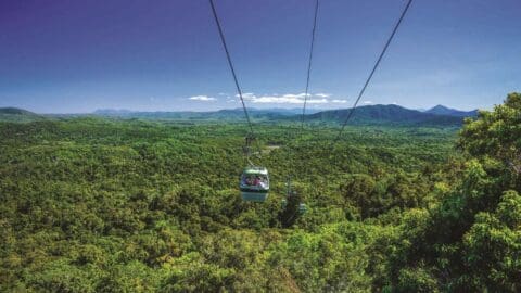 A cable car travels over a lush rainforest with a backdrop of distant mountains under a clear blue sky, offering an awe-inspiring view of nature's splendor.