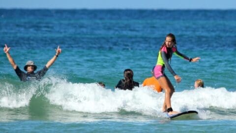 A person in a colorful wetsuit surfs a small wave while another in the background raises both arms in a peace sign gesture, promoting the spirit of ecotourism. Several people are visible enjoying the water behind them.