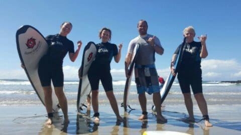 Four people stand on a beach with surfboards, making hand signs and smiling. They are wearing wetsuits and ankle leashes, enjoying the ocean waves behind them as part of their nature tour.