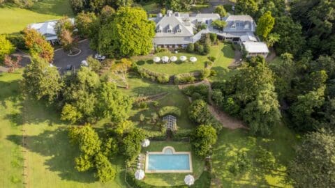 View from above on a generous property with a large white building, luscious garden and a separate pool area at the foot of the garden.