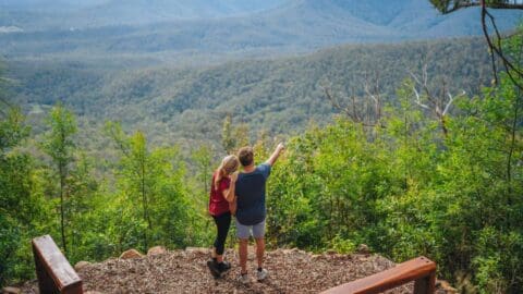 A couple standing on platform pointing in the distance while admiring the mountains covered in bushland.