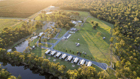 Aerial view of Splitters Farm showcases a rural agricultural landscape with farm buildings, green fields, a small lake, and dense forested areas under a setting sun.