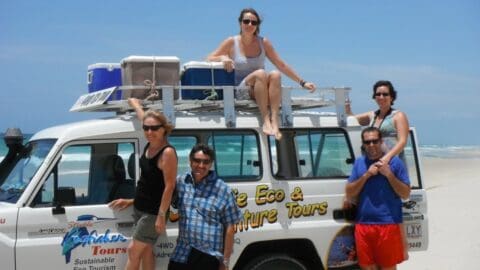 A group of five people pose on and around a white four-wheel-drive vehicle branded with "Eco & Adventure Tours" on a beach with ocean waves in the background, showcasing their dedication to ecotourism.