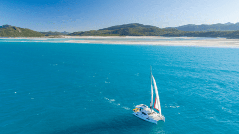 A sailboat gliding in the blue large ocean with a beautiful coast line with green hills and a white beach.