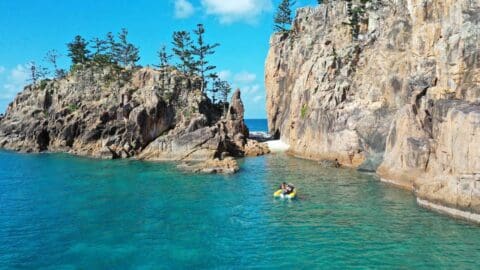 A person on a yellow dinghy floating in front of a large cliff and a rocky island.