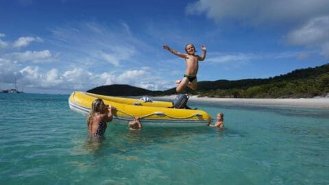 A family playing on a yellow dinghy in shallow water close to a white empty beach and green hills.