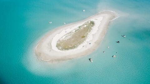 Aerial view of a small, sustainable sandbar island surrounded by turquoise water with boats anchored nearby.