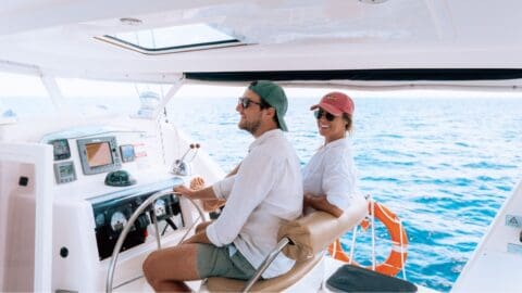 A man and woman are seated in the cockpit of a boat on the open sea. The man is steering while the woman smiles at the camera. Both are wearing casual clothing and caps, enjoying their sustainable maritime adventure.