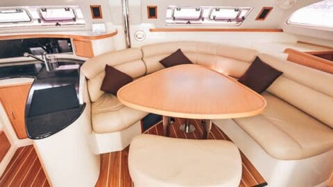 Interior of a boat with a curved beige leather seating area, triangular wooden table, and small black and wooden kitchen area designed for sustainable accommodation.