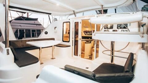 The interior of a boat showcases a dining area with a rectangular table and cushioned seating, leading to an open door to the cabin area. The steering station and kitchen are visible, reflecting sustainable design principles ideal for eco-conscious travelers.