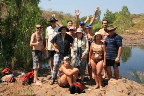 A group of people in casual clothing and swimsuits gather by a rocky, natural pool in an outdoor setting with trees and bushes. Some are standing while others squat, and one person raises their hands, seemingly celebrating the adventure provided by Tide Charters.