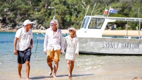 Three people walk barefoot along a beach next to a boat labeled "Sydney Harbour" with an Australian flag, trees, and a sandy shore in the background, enjoying sustainable ecotourism.