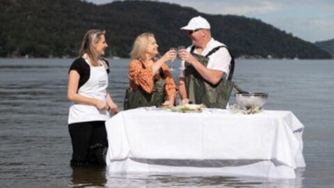 Three people stand in waist-deep water around a table with a white tablecloth. Two toast with wine glasses, while the third person stands beside them. Mountains are in the background, highlighting the beauty of nature and sustainable practices in ecotourism.