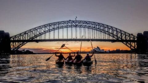 Four people are kayaking on water at sunset, with the silhouette of an arched bridge and a building with sail-like structures in the background, enjoying an ecotourism adventure.