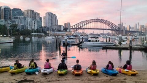 A group of people in kayaks listens to an instructor on a beach with a skyline and bridge in the background under a pink sunset sky, experiencing the serene beauty of nature.