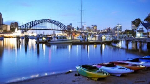 Sustainable kayaks rest on a sandy shore against a backdrop of a bridge and cityscape at dusk. Boats are docked nearby with reflections on the calm water, creating a serene scene that reflects ecotourism at its best.