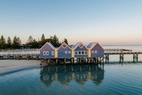 An image of a row of blue boathouses on stilts extending over calm water, with a pier and sandy shore visible. Trees are in the background, and the sky is clear, capturing the tranquil beauty often seen at Busselton Jetty.