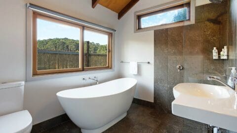 A modern bathroom with a freestanding bathtub, toilet, sink, and a walk-in shower. Large windows offer views of lush greenery outside, promoting sustainable living.