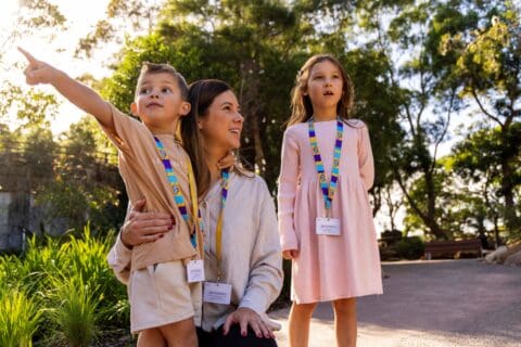 A woman kneels outdoors with two children, all wearing lanyards with badges. One child points excitedly to the distance while the other looks on with curiosity. Trees and a path are in the background, hinting at their adventure in Taronga's conservation area in Australia.