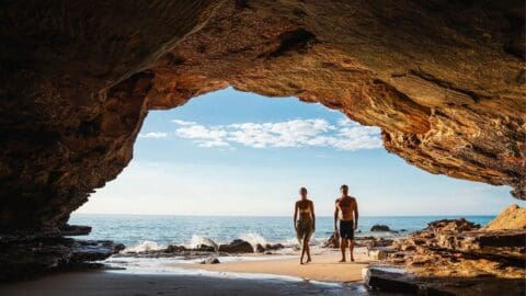 Two people walk on an eco-friendly resort's sandy beach framed by a rocky cave, with the ocean and a partly cloudy sky in the background.