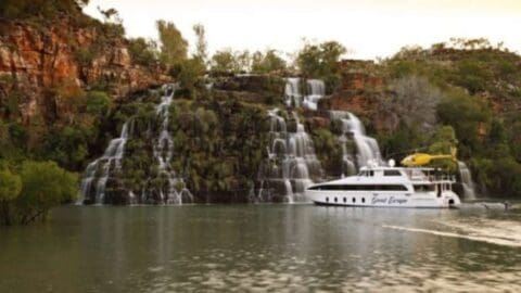 A white yacht is docked in front of a multi-tiered waterfall cascading down a rocky cliff into a river, surrounded by lush vegetation, creating the perfect setting for an ecotourism tour.