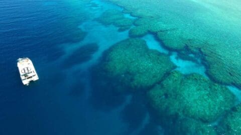 A boat is anchored in deep blue water near a vibrant, shallow coral reef visible beneath the surface, exemplifying sustainable ecotourism practices.