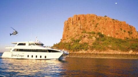 A white yacht named "Great Escape" on the water near a rocky island with greenery and a helicopter flying nearby under a clear sky, offering luxurious accommodation.