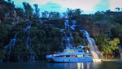 A white yacht named "Great Escape" is anchored in front of a multi-tiered waterfall surrounded by lush greenery and rocky terrain at dusk, offering luxurious accommodation amidst nature's beauty.