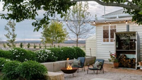 A fire pit with two chairs on a terrace surrounded by a garden and the wall of a white wooden house.