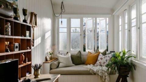 An inviting daybed nook with many cushions and a blanket surrounded by white wooden walls, windows and a book shelf.