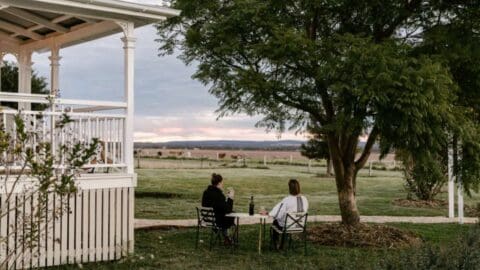 Two women sitting next to each other on a little table under a tree next to a white wooden house while overlooking the green plains in front of them.