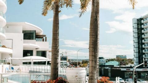 Two tall palm trees stand by the modern white architecture of The Sebel Maroochydore. In the background, more buildings and a partly cloudy sky are visible.