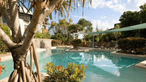Outdoor swimming pool with clear water, surrounded by trees and plants at The Sebel Twin Waters. Lounge chairs offer relaxation, while a shaded area with a green canopy provides comfort.