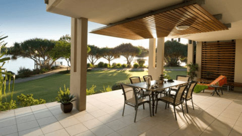 A covered patio with a dining table set and chairs at The Sebel Twin Waters overlooks a garden and a body of water. The area is surrounded by trees and lush greenery.