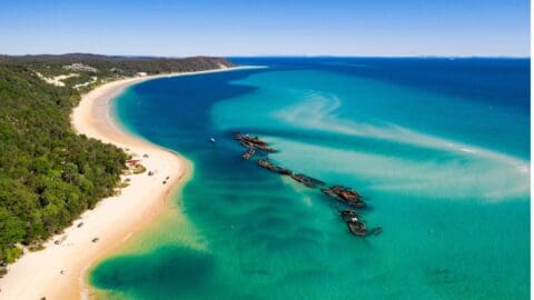 Aerial view of a beach with clear turquoise water, partially sunken shipwrecks near the shore, and dense greenery on the left, captured exquisitely by The Tour Collective.