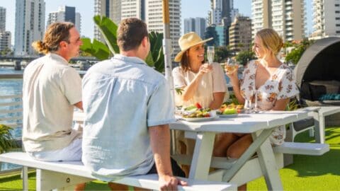 Four people are seated at an outdoor picnic table on a rooftop, surrounded by urban buildings, enjoying food and drinks—a perfect moment of collective travel laughter.