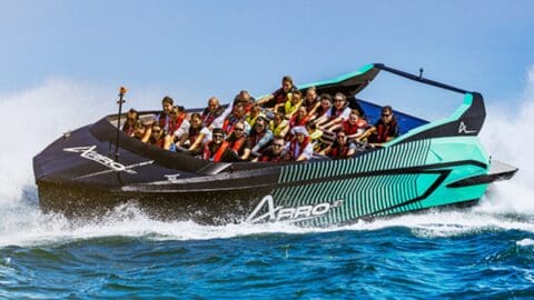 A Tour Collective speedboat packed with people wearing life vests cuts through the ocean, creating white splashes against the blue water.