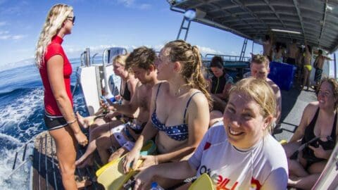 A group of people in swimwear are sitting on the edge of a boat, preparing for an ecotourism activity. A person in a red shirt is standing and talking to them. The boat is surrounded by water.