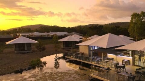 A serene resort with modern buildings under large, white, triangular shades is set against a sunset sky, with a reflective pond in the foreground and rolling hills in the background. This nature-inspired accommodation promises tranquility and comfort at every turn.