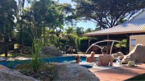 Three people relax in a small, outdoor pool surrounded by rocks and greenery. Two are adults, one is a child. A sun hat and bottled water are on the wooden deck. A shaded seating area is in the background, offering a perfect spot for those interested in ecotourism to unwind sustainably.