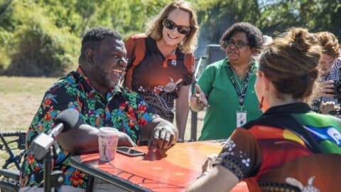 A group of people dressed in colorful shirts smile and engage in conversation outdoors, with one person seated and others standing around a table.