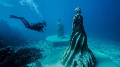 A scuba diver explores underwater sculptures on the ocean floor. The sculptures have abstract, textured designs and are mounted on platforms.