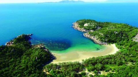 Aerial view of a coastal area with a small bay surrounded by green forest. Sandy beach curves around the bay, and clear blue-green water fills the bay. Distant landmasses are visible on the horizon.